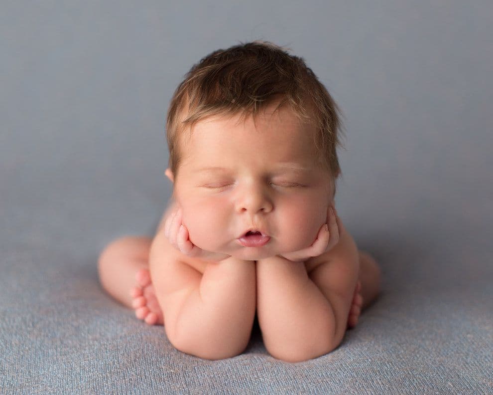 A sleeping newborn resting their face on their hands on a soft grey surface.